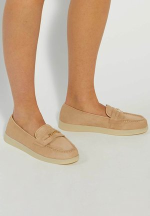 Beige suede loafers with a rounded toe, decorative stitching, a smooth texture, and a light-colored rubber sole. Gold-tone logo on the strap.
