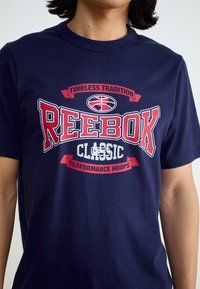Marineblå bomull t-skjorte med iøynefallende rød og hvit grafisk tekst med "REEBOK" og "CLASSIC PERFORMANCE HOOPS." Glatt tekstur, rund hals.