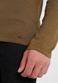 Olivegrünes Langarmshirt mit gerippten Ärmelbündchen, kombiniert mit dunkelblauen Jeans; mit einem kleinen metallischen Etikett am Saum.