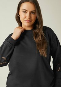 Svart sweatshirt med långa ärmar, prydd med intrikata utskärningar på muddar. Slät tygtextur, avslappnad passform, rund hals.
