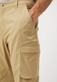 Khaki cargo bukser med avslappet passform, som har en sidelomme, glatt tekstur og knappelukking for sikker festing.
