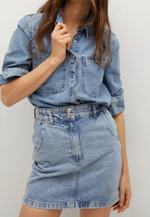 Kvinde iført en lyseblå denimskjorte med op rullede ærmer, stoppet ned i en matchende højtskåret denim mininederdel med dobbelt knaplukning.