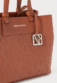 Ruskea teksturoitu olkalaukku, jossa on kohotettu "Armani Exchange" -kirjoitus, sileät yksityiskohdat, kaksoiset kantohihnat ja hopeinen logo-osa.