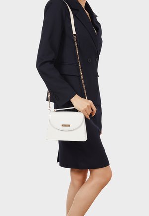 Donna con blazer nero e gonna che tiene una borsa bianca con tracolla a catena dorata su uno sfondo chiaro e semplice.