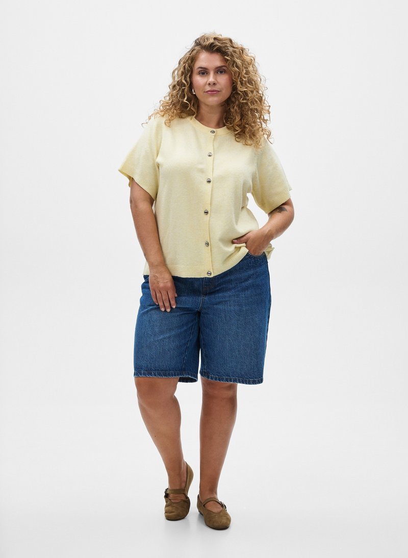 Frau mit lockigem Haar, die ein blassgelbes kurzärmeliges, geknopftes Oberteil, Jeans-Knielangshorts und braune flache Schuhe trägt, steht mit der Hand in der Tasche.