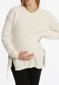 Eines cremefarbenen gestrickten Pullovers mit gerippter Textur, überschnittenen Schultern und seitlichen Bindebändern; kombiniert mit schwarzen Hosen. Chunky goldene Kettenhalskette.