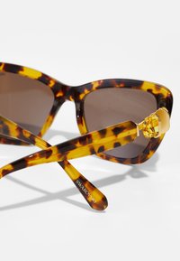 Ochelari de soare cu model țestoasă, lentile maro, rame groase și accente din cristal pe brațe. Logo-ul "Swarovski" pe un temp.