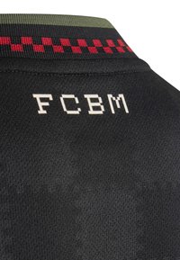 Zwarte jersey stof met een gestructureerd patroon; heeft een groen en rood geruit streep op de kraag; "FCBM" in het wit gedrukt op de achterkant.
