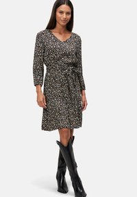 Robe florale noire avec un décolleté en V, des manches longues et une ceinture à la taille. Fabriquée en tissu doux, elle présente une jupe évasée avec des accents blancs et jaunes. Bottes noires jusqu'aux genoux.