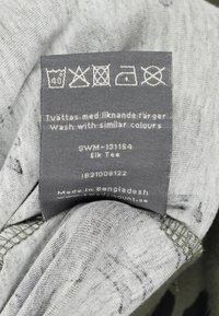 Gray fabric clothing label featuring care instructions, text, and symbols. Tag displays "Elk Tees," product code SWM-131154, made in Bangladesh.
