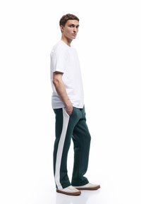 Hvid bomulds t-shirt parret med grønne sweatpants med hvide sidelister. Modellen har hvide sko med tan accenter på og hænderne i lommerne.