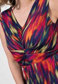 Värikäs hihaton Mekko, jossa on V-mallinen kaula-aukko ja vyötäröllä käänteentekevä yksityiskohta. Kankaan pinnassa on poimutettu rakenne, jossa on eloisia, kiemurtelevia kuvioita.