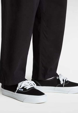 Sneakers basse in tela nera con suole e lacci bianchi in gomma, indossate con pantaloni neri a gamba dritta su sfondo bianco.