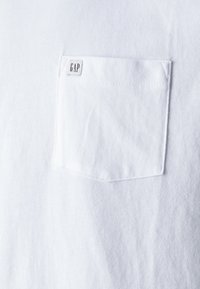 T-shirt en coton blanc avec une poche sur la poitrine gauche, présentant une petite étiquette "GAP" cousue dans la poche. Texture douce et design simple.