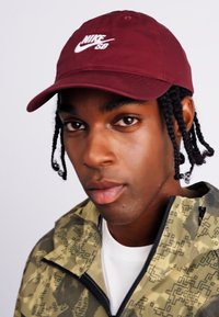Căciulă Nike SB de culoare bordo cu logo brodat, purtată cu o jachetă de culoare verde măsliniu, cu modele geometrice. Textură netedă a pielii, păr împletit.