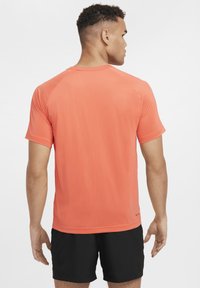 Kortermet atletisk t-skjorte i klar oransje stoff, med mesh tekstur, rund hals og en figursydd design. Båret sammen med svarte shorts.
