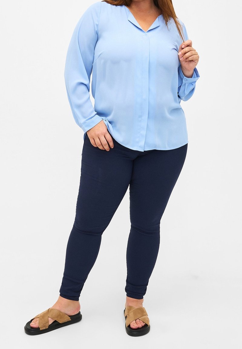 Femme portant un chemisier bleu clair à manches longues, un pantalon bleu foncé ajusté et des sandales beige, debout devant un fond blanc uni.