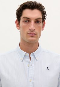 Camicia a bottoni a righe blu chiaro con colletto, presenta un piccolo logo ricamato di teschio e ossa incrociate sul petto. Materiale in cotone.