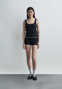 Νεαρή γυναίκα στέκεται μπροστά σε απλό τοίχο φορώντας μαύρο τοπ χωρίς μανίκια και μαύρα σορτς με λευκή ρίγα, λευκές κάλτσες και μαύρα παπούτσια Mary Jane.