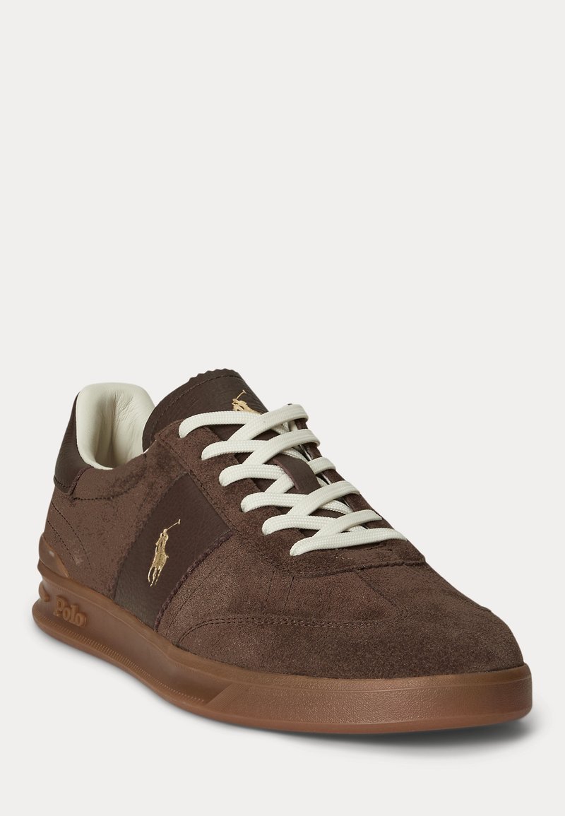 Baskets en daim marron avec lacets blancs, semelle en caoutchouc et logo Polo doré sur le côté et la languette, présentées sous un léger angle sur fond blanc.