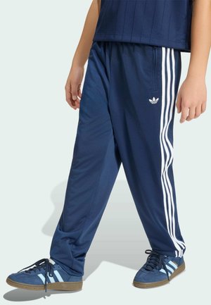 Person wearing navy blue Adidas track pants with white stripes and matching Adidas sneakers standing against light background.
