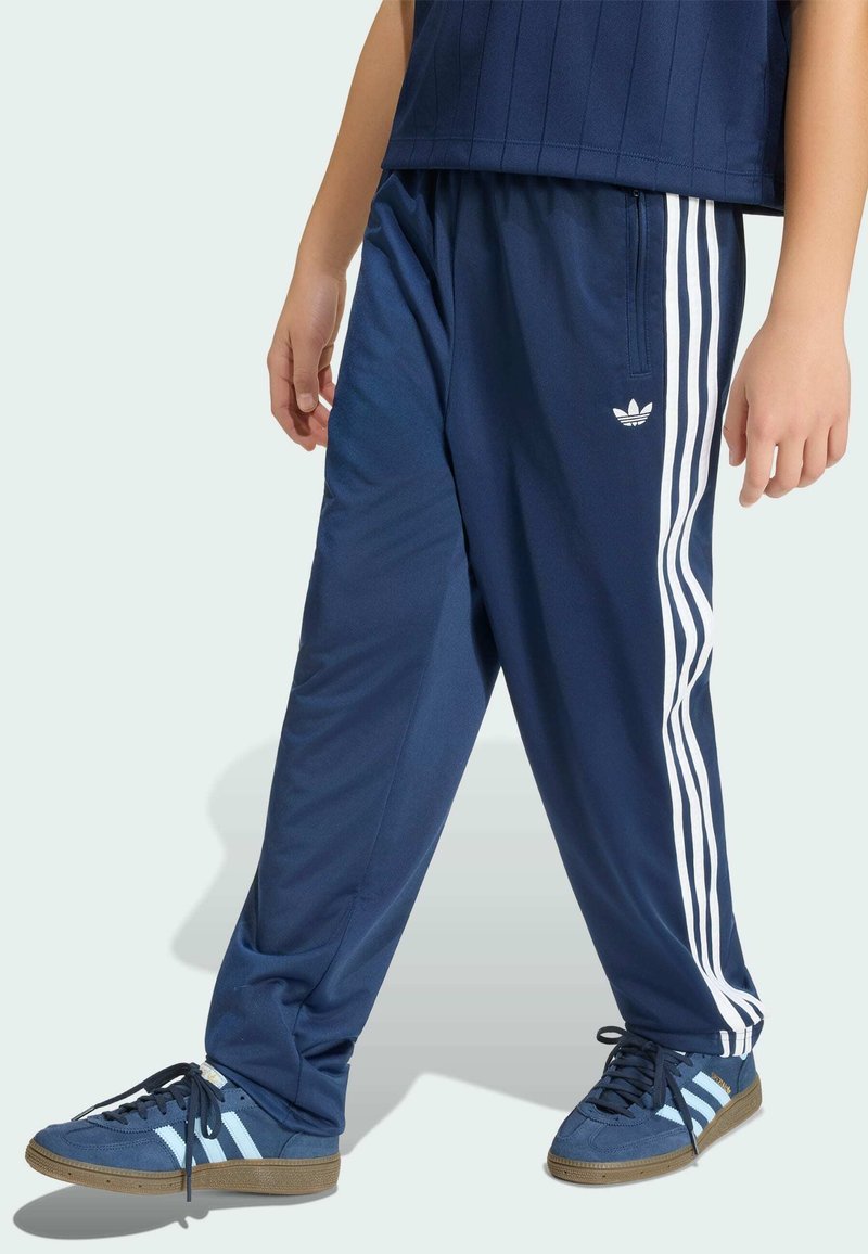 Άτομο που φοράει σκούρο μπλε φόρμα Adidas με λευκές ρίγες και αντίστοιχα αθλητικά παπούτσια Adidas, στέκεται μπροστά σε φωτεινό φόντο.