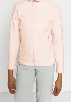 Person wearing a light pink zip-up fleece jacket with pockets and light gray pants, standing against a plain background.