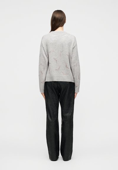 Γυναίκα με μακριά καστανά μαλλιά, φορώντας ανοιχτό γκρι πουλόβερ με σχέδιο και μαύρο υφασμάτινο παντελόνι με φαρδιά μπατζάκια, στέκεται γυρισμένη πλάτη σε λευκό φόντο.