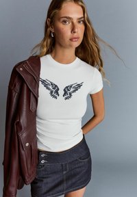Young woman with long hair wears white T-shirt with black wing print, dark denim skirt, and holds maroon leather jacket over shoulder.