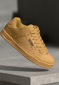 Yellow skate shoe with a textured upper, featuring prominent embossed branding and structured design. Thick laces and cushioned tongue.