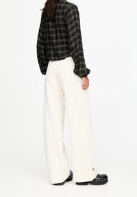 Camicia cropped a quadri nera e verde con maniche lunghe, abbinata a pantaloni bianchi larghi e zoccoli neri con borchie di metallo argentato.