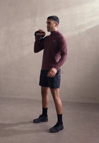 Sportaš drži crni kettlebell, nosi tamnocrvenu majicu dugih rukava i crne šortseve, sa crnim čarapama i sportskim cipelama na sivoj podlozi.