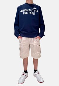 Felpa navy con testo bianco "Aeronautica Militare", pantaloni cargo beige, sneakers bianche con accenti rossi e calzini neri.