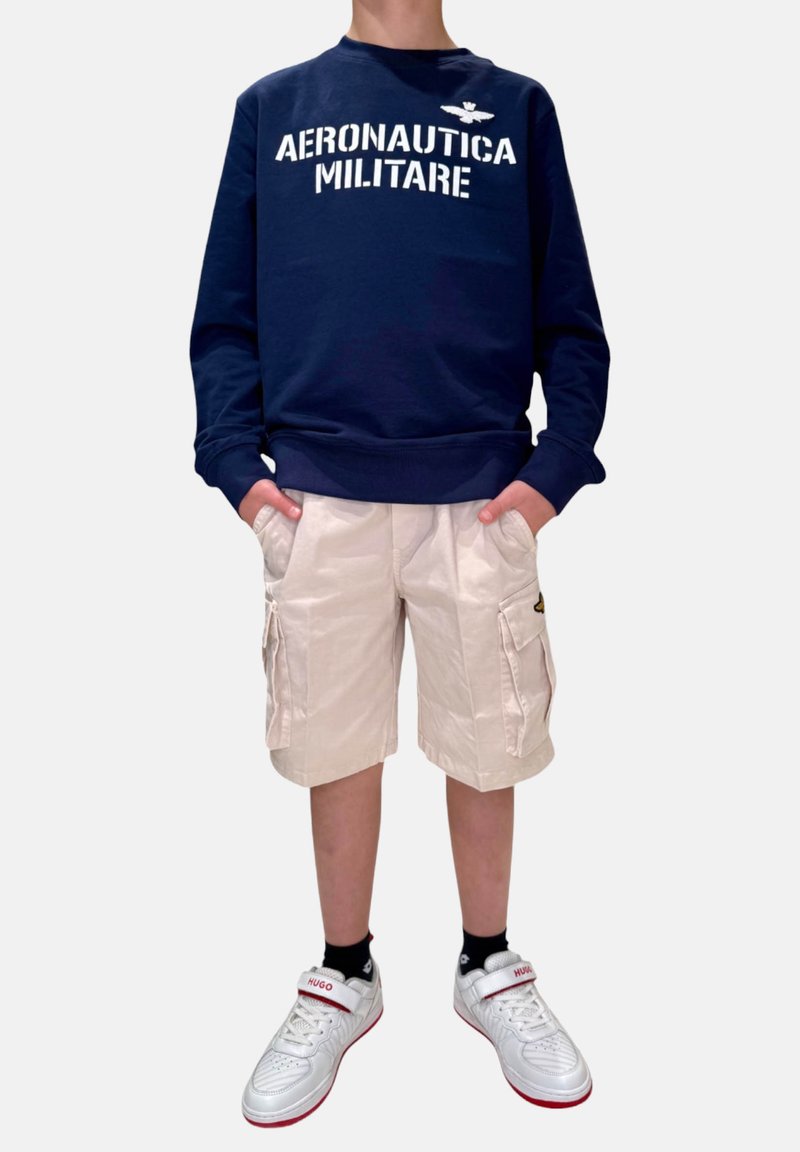 Felpa navy con testo bianco "Aeronautica Militare", pantaloni cargo beige, sneakers bianche con accenti rossi e calzini neri.