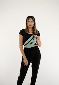 Marsupio verde a fantasia geometrica con cerniera nera e logo, indossato a tracolla; abbinato a pantaloni neri a vita alta e a una maglietta nera.