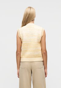 Ξανθιά γυναίκα με ίσια μαλλιά φοράει αμάνικη κίτρινη και λευκή πλεκτή μπλούζα και μπεζ παντελόνι ψηλόμεσο, γυρισμένη με την πλάτη προς ένα απλό φόντο.