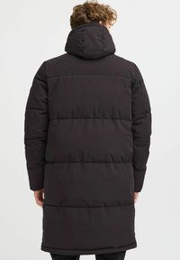 Melnā pūkainā jaka ar kapuci, kurai ir quilted dizains un garas piedurknes, beidzas zem ceļgaliem. Taisna apakšmala un teksturēta virsma.