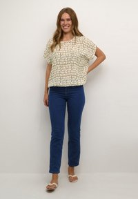 Cremefarvet top med et struktureret rutemønster, parret med tætsiddende blå jeans og lyse sandaler. Outfittet er afslappet og moderne i stil.