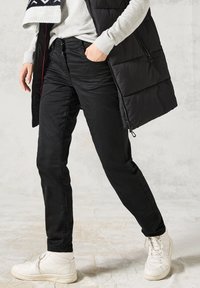 Svarta, slim-fit byxor med en slät yta och subtil lyster. Matchas med en ljusgrå tröja och en puffy svart väst. Vita hög-top sneakers.