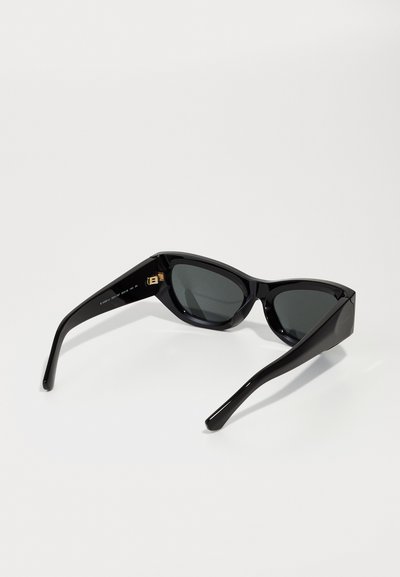 Óculos de sol pretos em forma de gato com lentes escuras, armação de plástico brilhante, ângulos acentuados e hastes afiladas. Detalhe de hardware notável perto da articulação.