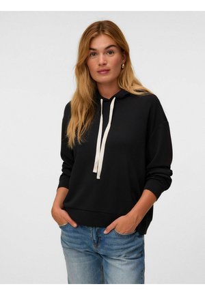 Donna con lunghi capelli biondi che indossa una felpa nera con cordini bianchi e jeans blu, in piedi con le mani nelle tasche davanti a uno sfondo semplice.