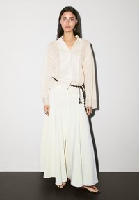Chemise beige clair à boutons surdimensionnée associée à une longue jupe maxi crème. Ceinture tressée remarquable et sandales plates simples.