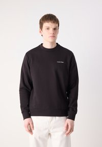 Fekete környakú pulóver, amely pamut anyagból készült, diszkrét fehér Calvin Klein logóval, hosszú ujjakkal és texturált felülettel.