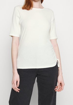 Frau trägt ein weißes, geripptes Kurzarmoberteil mit seitlichem Kordelzugdetail und schwarze Hosen vor einfarbigem Hintergrund.