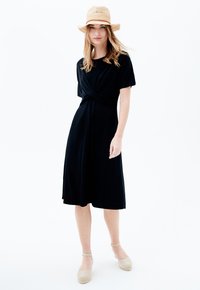 Robe noire au genou avec un design à l'avant torsadé, manches courtes et coupe décontractée. Associée à des chaussures espadrilles beiges et un chapeau en paille.