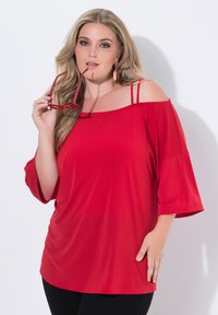 Rode off-shoulder top met korte, brede mouwen, voorzien van dubbele banden. Zacht, gladde stof met een ontspannen pasvorm. Zwarte broek.