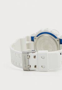Montre G-SHOCK avec un bracelet en résine blanche et des éléments en métal argenté. Comprend une accentuation bleue et un dos texturé avec un boîtier circulaire.