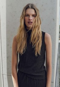 Black sleeveless top with a textured fabric, featuring a high neckline and subtle pleats. Model has long, straight hair.