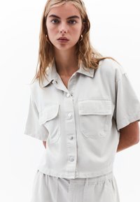 Jasnoszara, krótkosleeva koszula z guzikiem z przodu, dwoma kieszeniami na piersi i wywijanym kołnierzykiem. Gładka tkanina o luźnym kroju.