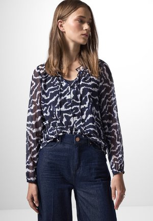 Blusa navy trasparente con un motivo astratto bianco, con scollatura arricciata e maniche lunghe, abbinata a jeans in denim scuri a vita alta.